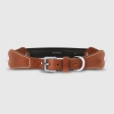 A dog collar in smooth, natural bovine leather, handmade in a Polish artisan workshop. Designed by the Italian designer Francesco Rigucci, it is recognisable by its signature shape and the jewellery-like metal studs carrying the brand logo. The outer layer is caramel vegetable-tanned bovine leather with contrasting beige stitching, over soft padding and a chocolate leather lining. The D-ring, buckle and logo studs are solid brass in silver finish, nickel-free and protected with a durable polished coating. Adjustable length – check the measurements to choose the right size for your dog. Available on its own or as a set with the matching leather leash and lambskin gloves.