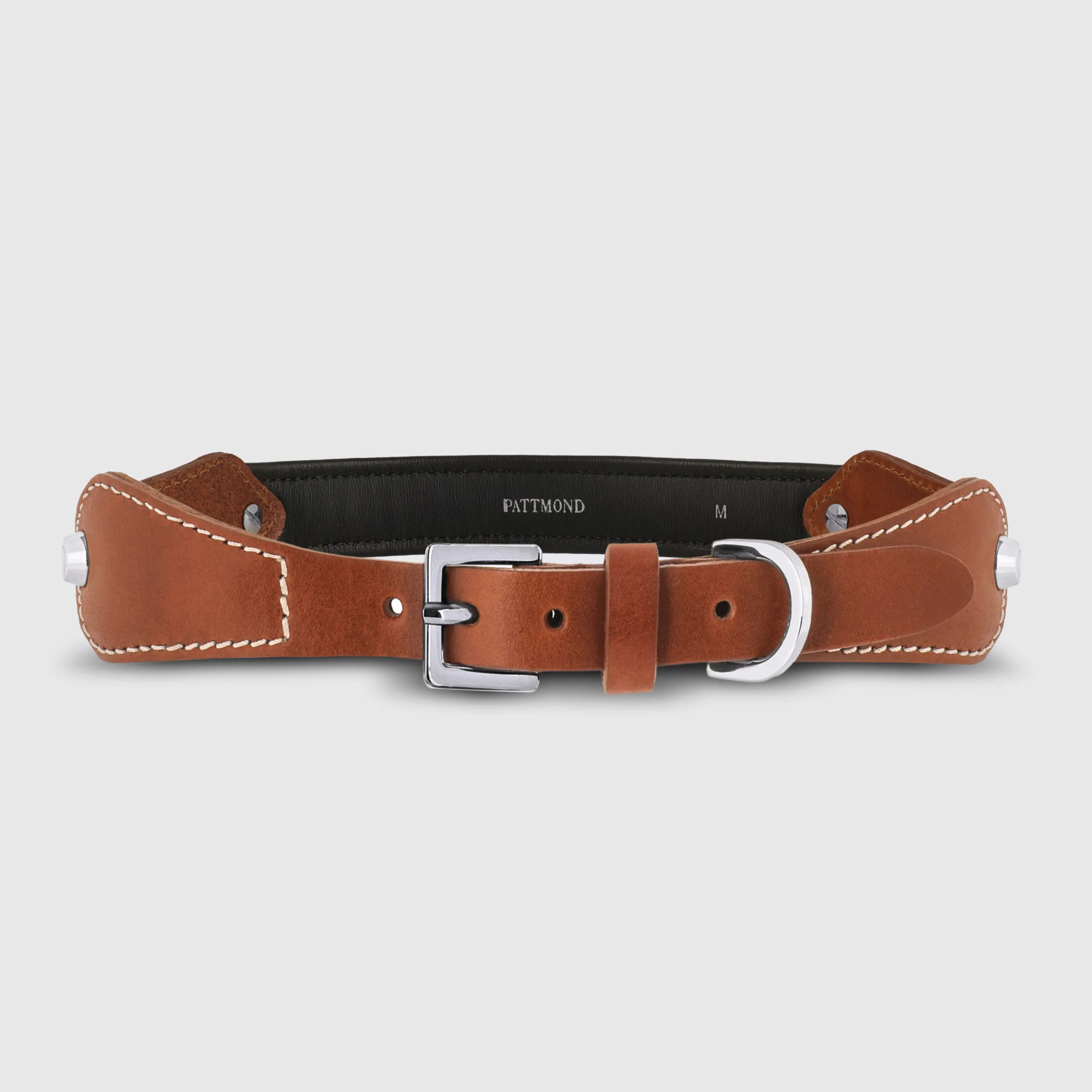 A dog collar in smooth, natural bovine leather, handmade in a Polish artisan workshop. Designed by the Italian designer Francesco Rigucci, it is recognisable by its signature shape and the jewellery-like metal studs carrying the brand logo. The outer layer is caramel vegetable-tanned bovine leather with contrasting beige stitching, over soft padding and a chocolate leather lining. The D-ring, buckle and logo studs are solid brass in silver finish, nickel-free and protected with a durable polished coating. Adjustable length – check the measurements to choose the right size for your dog. Available on its own or as a set with the matching leather leash and lambskin gloves.