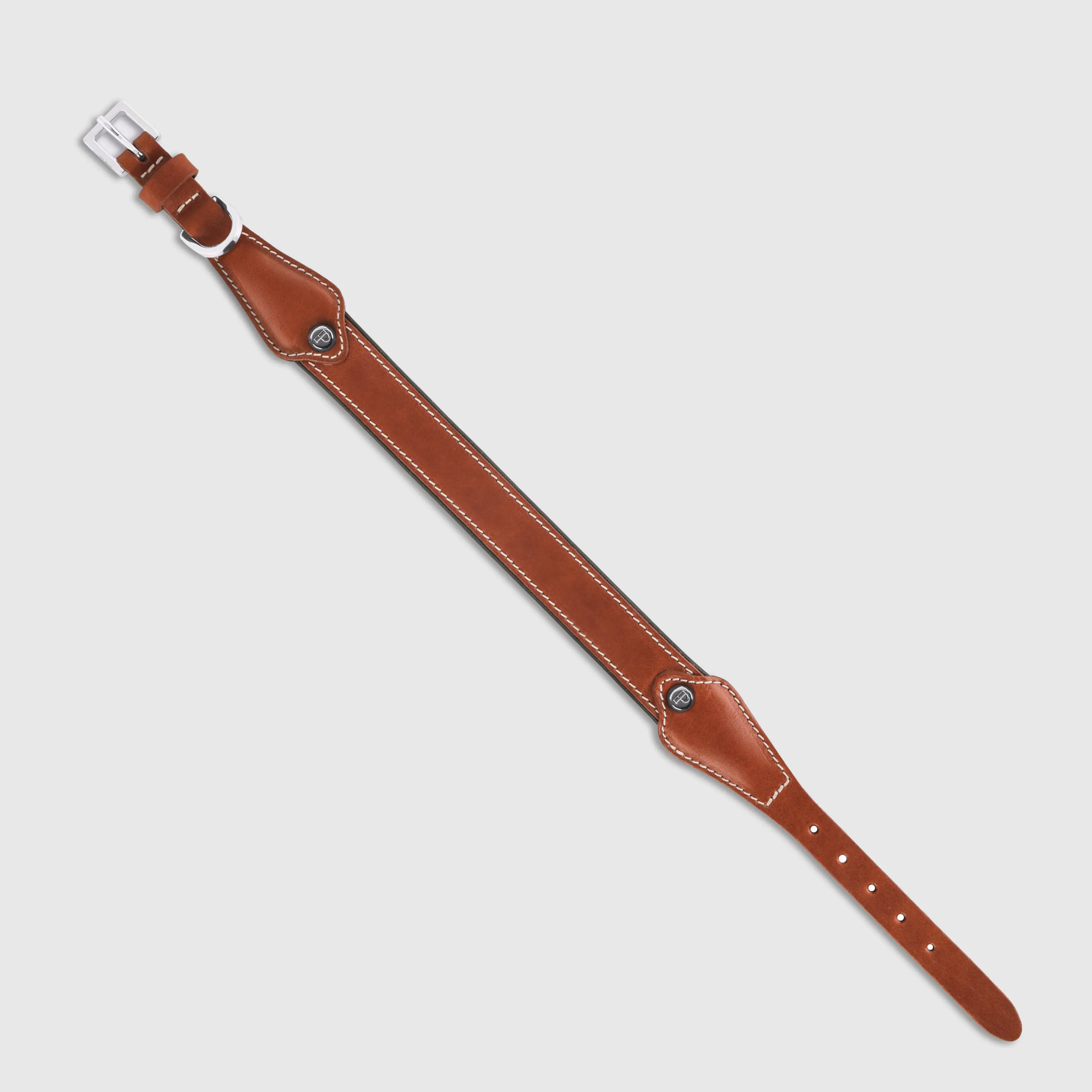 A dog collar in smooth, natural bovine leather, handmade in a Polish artisan workshop. Designed by the Italian designer Francesco Rigucci, it is recognisable by its signature shape and the jewellery-like metal studs carrying the brand logo. The outer layer is caramel vegetable-tanned bovine leather with contrasting beige stitching, over soft padding and a chocolate leather lining. The D-ring, buckle and logo studs are solid brass in silver finish, nickel-free and protected with a durable polished coating. Adjustable length – check the measurements to choose the right size for your dog. Available on its own or as a set with the matching leather leash and lambskin gloves.